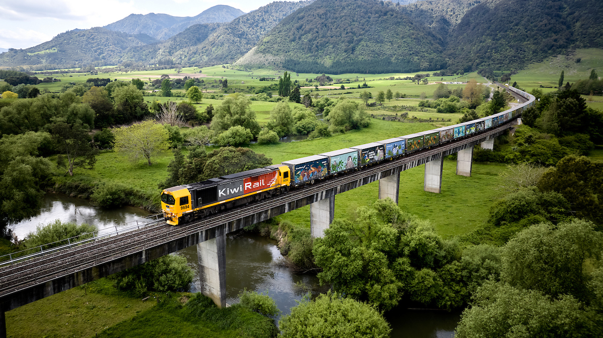 A KiwiRail DL locomotive hauling containers across a river viaduct in green hill country